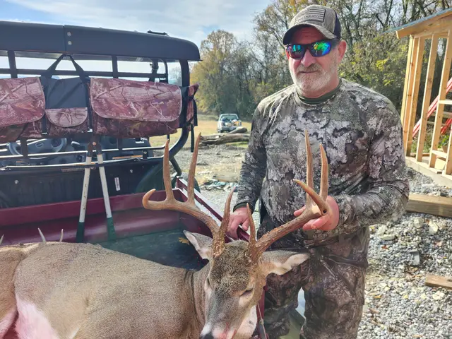 Hunter in camouflage and mirrored sunglasses holding a buck’s antlers beside a utility vehicle.