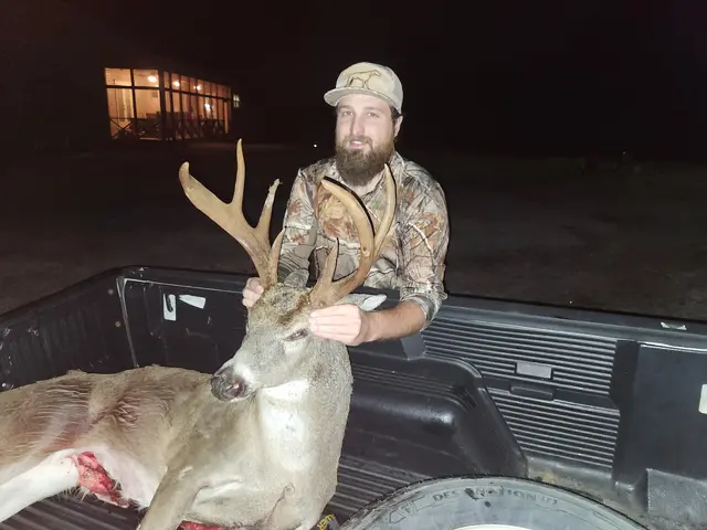 Hunter in a camouflage shirt and cap posing with a buck in a pickup truck bed at night.