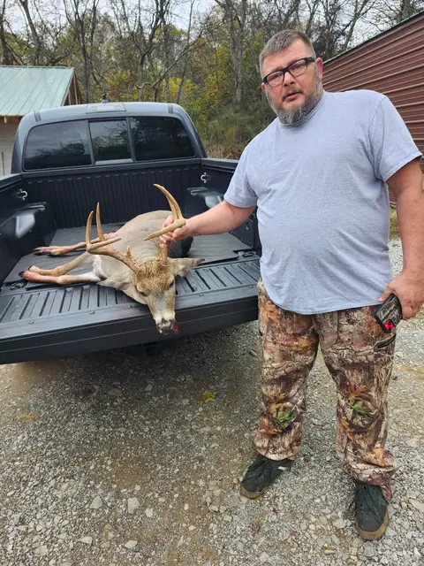 Hunter in a gray shirt standing beside a buck on an open pickup truck tailgate.