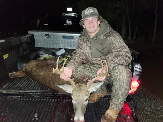 Hunter in camouflage kneeling beside a buck in a white pickup truck bed at night.