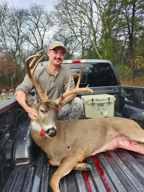 Hunter in a cap and camouflage holding a buck’s antlers in a pickup truck bed outdoors.