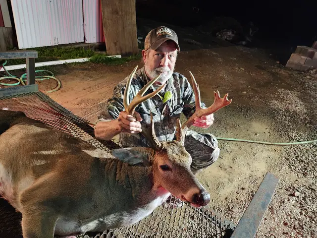 Hunter in camouflage holding a buck’s antlers beside a building at night.