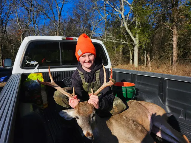 Hunter wearing a bright orange knit cap posing with a buck in a pickup truck bed in daylight.