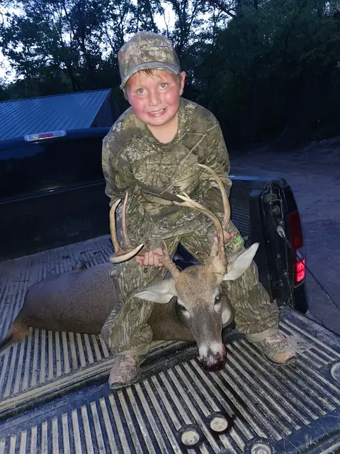 Young hunter in camouflage and a cap posing beside a buck in a pickup truck at night.