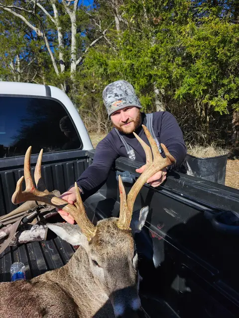 Young hunter wearing a knit cap and camouflage posing with a buck in a pickup truck bed.